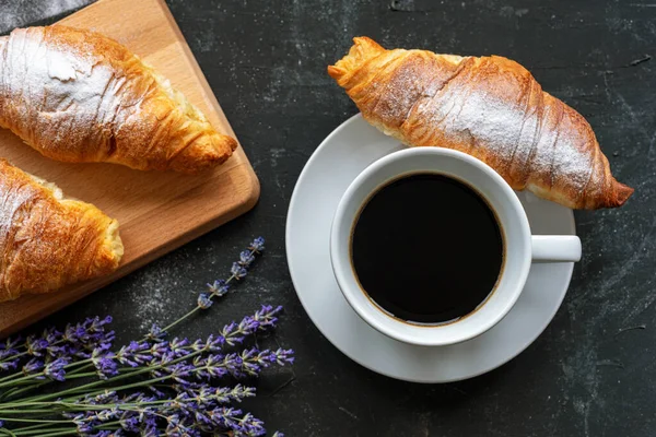 A cup of coffee and croissants, bunch of lavender flowers on black table, top view. Morning breakfast concept