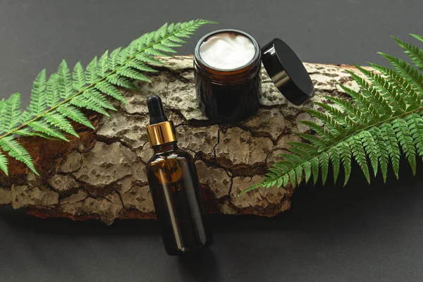 Cosmetic bottle and cream jar on black background with wood and greenery. Natural cosmetics concept. Top view