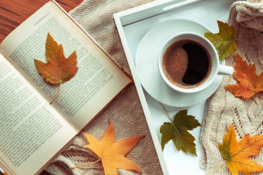 Sonbahar yapraklı bir tepside bir fincan kahve, bir kitap ve bir örgü süveter. Konforlu ev, sonbahar konsepti. Üst manzara, düz uzanma.