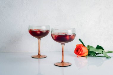 Beyaz zemin üzerinde buz küpleri ve turuncu gül olan iki zarif kırmızı kokteyl bardağı. Yakın çekim..