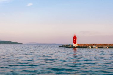Kayalık sahilde duran kırmızı deniz feneri. Arka planda sakin deniz ve günbatımı gökyüzü var. Krk Adası, Hırvatistan.