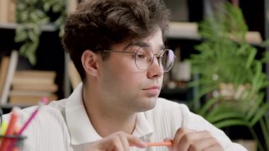 Concentrated and focused student learning online on a website. Young man with eyeglasses working remotely at home office, taking notes in work documents and looking at laptop typing on laptop
