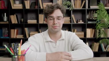 A boy student sitting on a bench in a classroom looks directly at the camera, smiles and demonstratively raises his hand up as a sign that he wants to answer,to give his opinion.Good manners at school