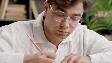Young specialist attends an online advanced course taking notes with a pencil in a notebook. Boy with glasses and a classic shirt attends an international conference for a job