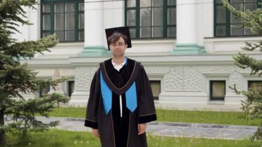 Proud graduate in cap and black robe walks confidently outside in the courtyard of the university campus and enjoys his achievements. Boy with glasses at the university graduation ceremony