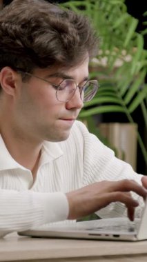 Genç bilişim uzmanı dikkatle yazıyor ve dizüstü bilgisayardaki klavyeye odaklanmış durumda. Gözlüklü çocuk modern bir ofiste çalışıyor ve web siteleri kuruyor. İş yerindeki sevimli çocuk.