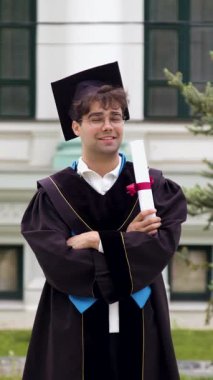 Siyah bornoz giymiş ve başında şapkası olan mezun bir çocuk elleri kollarının altında uluslararası bir üniversiteden mezuniyet diplomasını gururla tutuyor. Başarılı mutlu öğrenci.