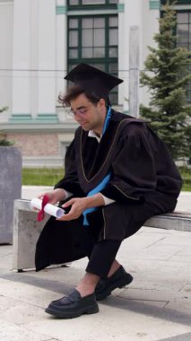 Young graduate in academic cap and gown rests happily and proudly holding his higher education diploma in his hand on a bench near the campus. The man dreams of future career successes.