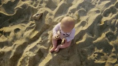 Boy is playing in soft sand on a beach, holding a small rock, with gentle waves and a bright sky creating a joyful atmosphere of childhood adventure