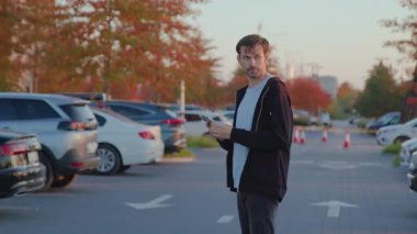Casual-dressed young man stands in a parking lot, holding smartphone, surrounded by parked vehicles, illustrating modern carsharing accessibility and urban mobility