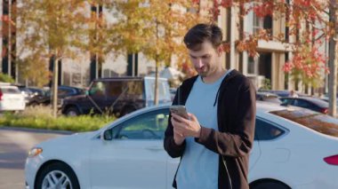 Erkek kullanıcı akıllı telefonda araba paylaşım uygulaması ile etkileşime girer, canlı şehir ortamında park halindeki arabanın yanında durur, kolaylık ve yenilik vurgulanır