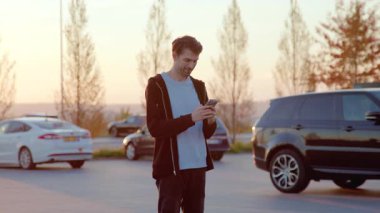 Şahıs, araçlarla çevrili bir otoparktaki araba paylaşım servisine ulaşmak için akıllı telefonuyla etkileşime girer ve gün batımının ılık ışığıyla aydınlatılır.