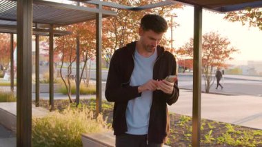 Sıradan giyinmiş bir adam, ağaçların ve çağdaş yapıların oluşturduğu canlı bir alışveriş bölgesindeki akıllı telefonlara odaklanmış durumda.