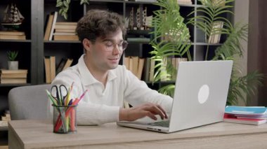 Kıvırcık saçlı ve görme bozukluğu olan genç bir öğrenci hafifçe gülümseyip bilgisayar başında çalışıyor. Samimi bir ofisteki dizüstü bilgisayarda tatmin olmuş çocuk tipleri ve uzaktan internetten öğrenim görüyor.