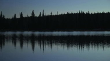 Siluetli ağaçlar sabahın erken saatlerinde camsı göle yansıyor..