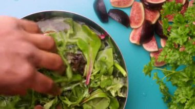A person prepares a vibrant green salad, mixing leafy greens and sliced figs.