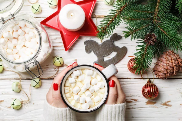 Hot chocolate (coffee, cocoa) with marshmallow in white Cup in women\'s hands. Christmas. New year. Fir branches, candles, cones, jingle bells and toys. Holiday. Selective focus, top view