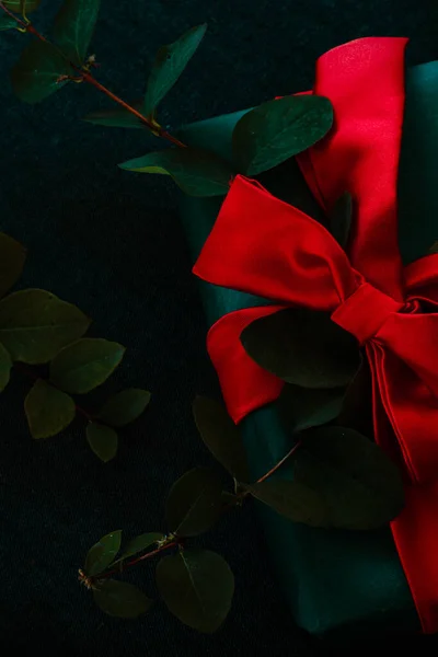 Close-up van de kerstgeschenkdoos bovenop. Groene geschenkdoos met een rode zijden strik op een donkere achtergrond, twijgen met groene bladeren — Stockfoto