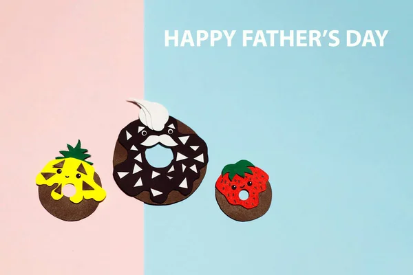 The family of chocolate donuts covered with different glazes was cut out of colored paper on a pink and blue background for your Father\'s Day design. There is a place for text.