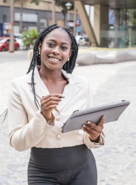 Afro-Amerikalı iş kadını bir iş ortamında tablet üzerinde çalışırken gülümsüyor. Dikey fotoğraf
