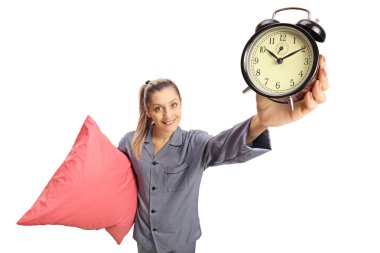 Young woman in pajamas holding a pillow and an alarm clock isolated on white background clipart