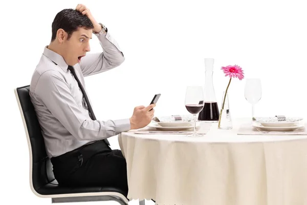 白い背景に分離されたレストランのテーブルに着席している彼の携帯電話を見てショックを受けた若い男 — ストック写真