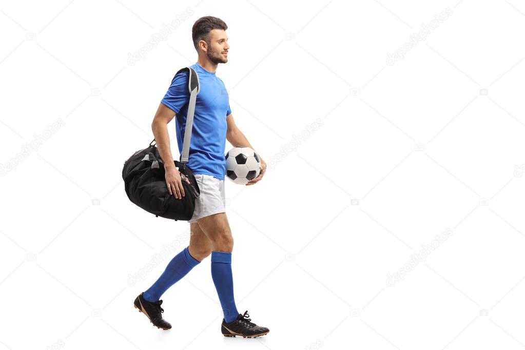 Full length profile shot of a soccer player with a bag and a football walking isolated on white background