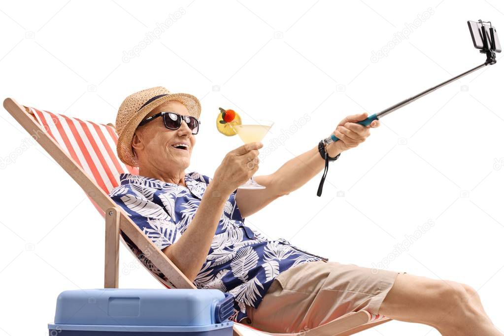 Elderly tourist with a cocktail sitting in a deck chair and taking a selfie with a stick isolated on white background