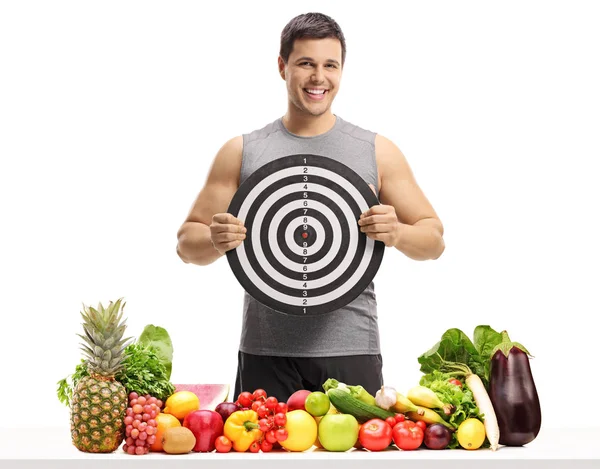 在一张桌子后面有水果和蔬菜的年轻人拿着一个在白色背景下被隔离的目标 — 图库照片