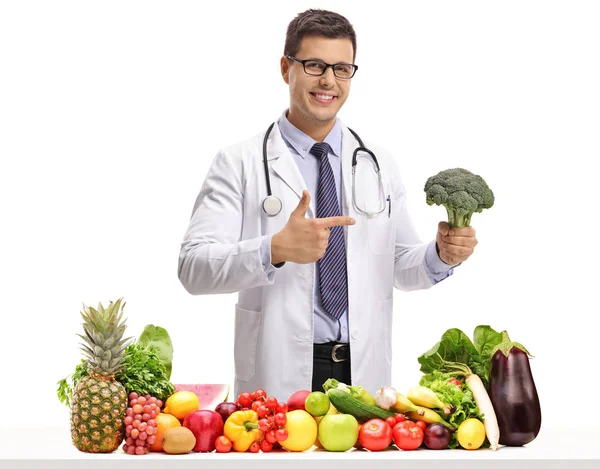 医生与花椰菜指着桌子后面的水果和蔬菜孤立的白色背景 — 图库照片