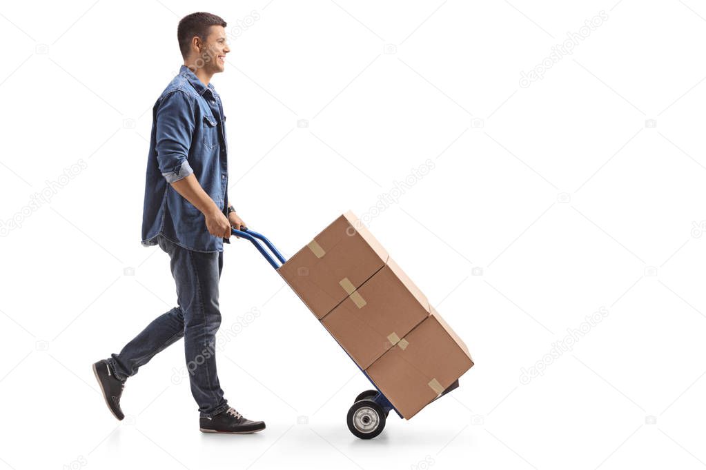 Full length profile shot of a young man pushing a hand truck with boxes isolated on white background