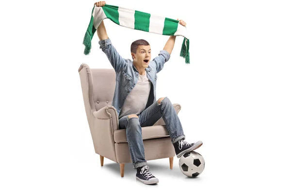 Overjoyed teenage soccer fan with a scarf and a football sitting in an armchair isolated on white background
