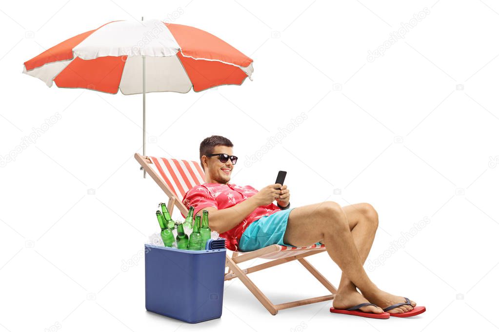 Tourist with a phone sitting in a deck chair with an umbrella next to a cooling box isolated on white background