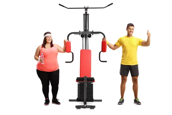 一个超重的妇女和男性健身教练的全长肖像站在一台运动机器旁边 在白色背景上显示拇指 — 图库照片