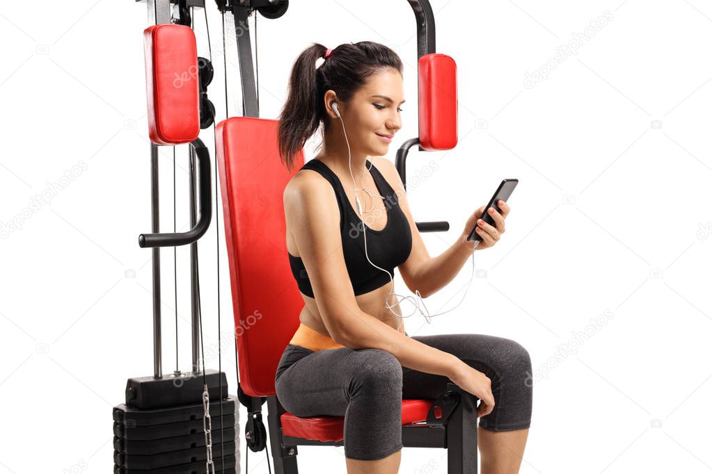 Young female with a cell phone sitting on an exercise machine isolated on white background