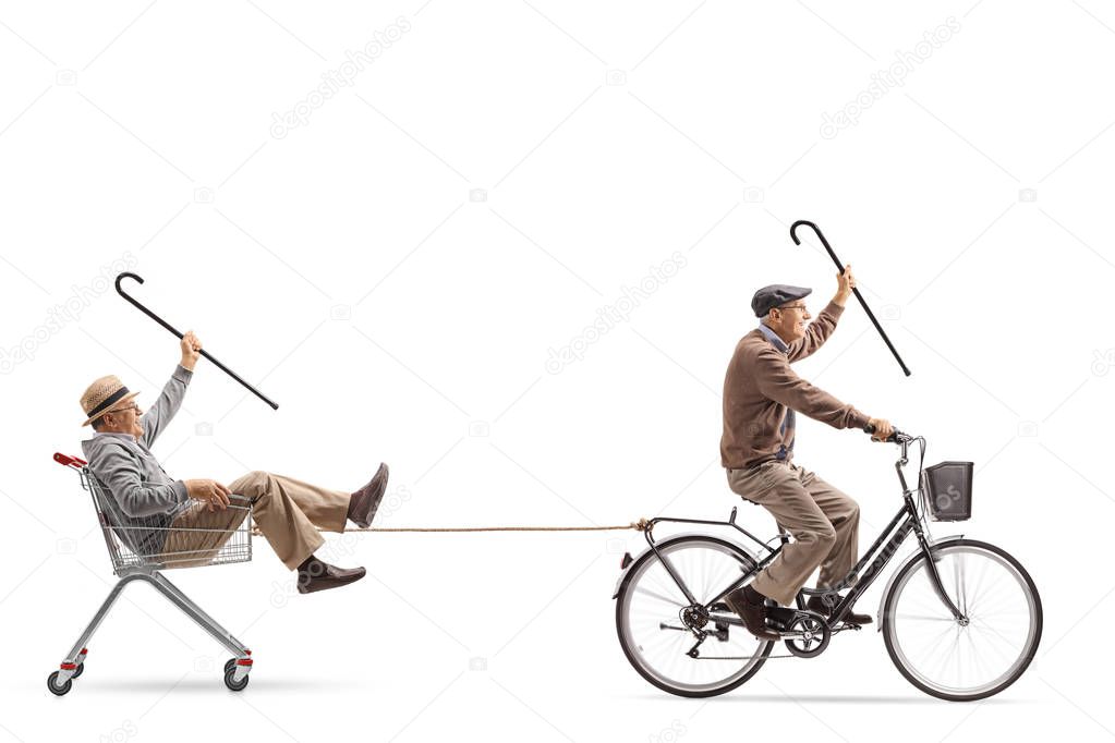 Senior riding a bicycle and pulling another senior inside a shopping cart with a rope isolated on white background