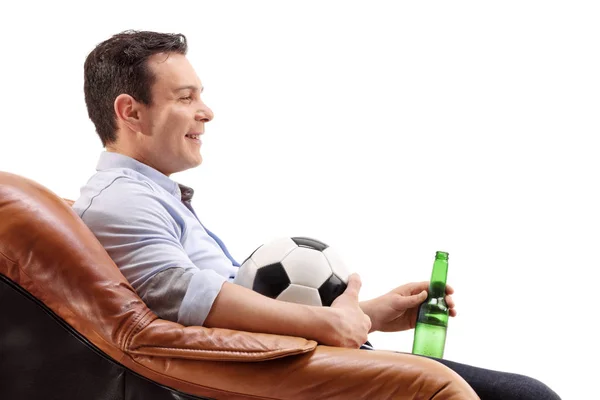 年轻男子与啤酒瓶和足球坐在扶手椅上孤立的白色背景 — 图库照片