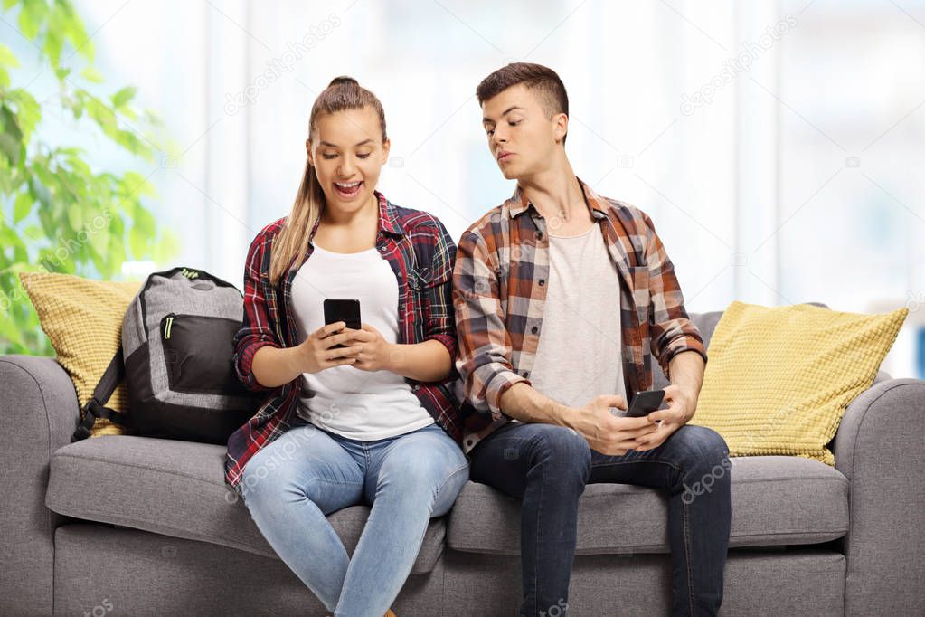 Teenage boy peeking at the phone of a teenage girl sitting next to him on a sofa