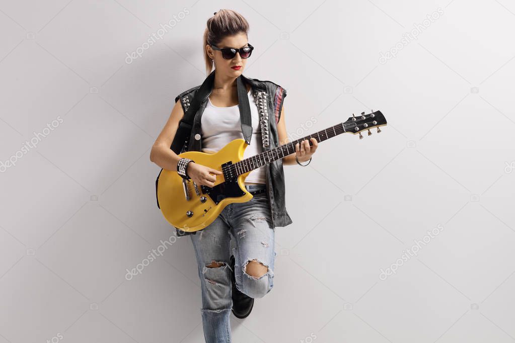 Female punker in a leather vest leaning on a wall and playing a guitar 