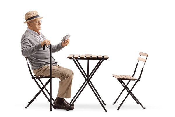 Full length shot of a senior man with a cane sitting at a table and playing cards alone isolated on white background