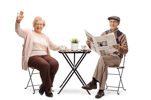 コーヒーのカップをテーブルに座って 手を振っている年配の女性の白い背景に分離された新聞を読む老人完全な長さの肖像画 — ストック写真