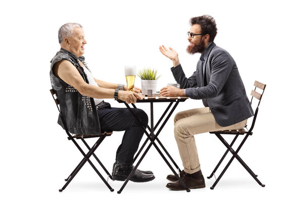 Full length profile shot of a mature man in a leather vest drinking beer at a table and talking with a young bearded man isolated on white background