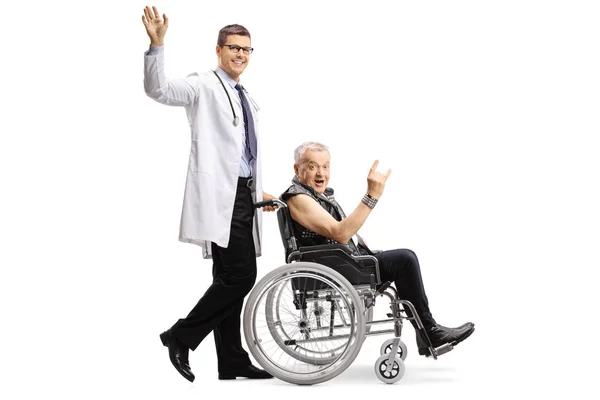 手を振って 白い背景で隔離車椅子サインイン ロックン ロールをもうけて成熟した男性患者を押す若い男性医師の全身ショット — ストック写真