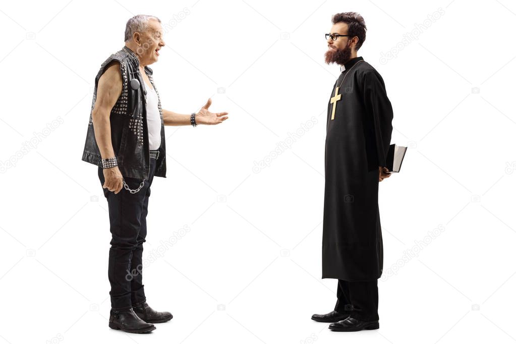 Full length profile shot of a mature male punker in a leather vest talking to a priest isolated on white background