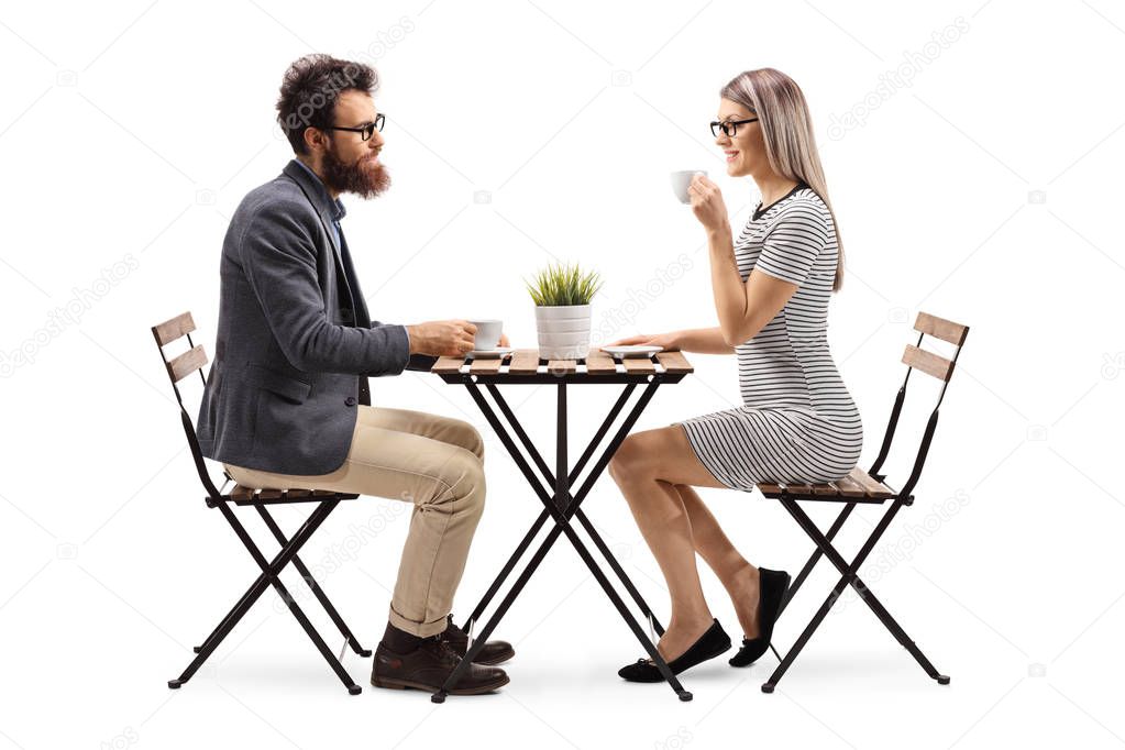 Full length profile shot of a young man and woman drinking coffee at a table isolated on white background