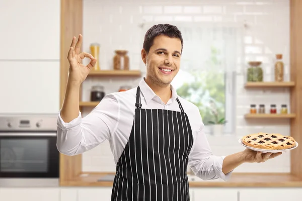 微笑的年轻人与围裙拿着馅饼在厨房里 用手做手势 — 图库照片