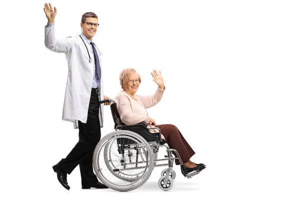 プロファイルショットを振るとの分離の白い背景を振って車椅子で年配の女性を押す若い男性医師の完全な長さ — ストック写真