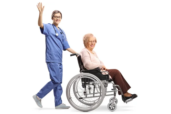 全長プロファイルショットを振るとの分離の白い背景を振って車椅子で年配の女性を押す若い女性看護師の — ストック写真