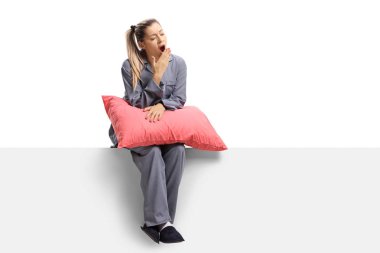 Full length shot of a young sleepy woman sitting on a panel with a pillow and yawning isolated on white background clipart