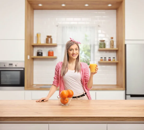 モダンなキッチンの木製カウンターの後ろに新鮮なオレンジ ジュースのガラスを持つ若い女性を笑顔 — ストック写真